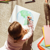 Top View: Little Girl Drawing Our Beautiful Green Planet Earth. Child Having Fun at Home on the Floor, Imagining Our Planet as a Happy Place with Clean, Sustainable Living. Cozy Sunny Day.